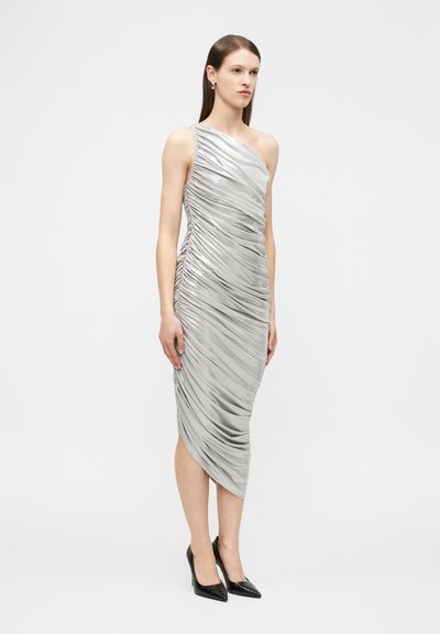 Γυναίκα που στέκεται φορώντας ένα ασημένιο μίντι φόρεμα ενός ώμου με πιέτες, συνδυασμένο με μαύρα μυτερά ψηλοτάκουνα, σε απλό φόντο.