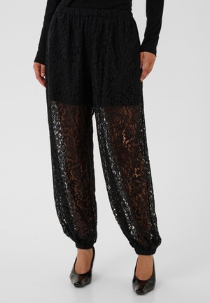Pantalon en dentelle noire avec taille et poignets élastiques, porté sur un short noir, associé à un haut noir à manches longues et des chaussures à talons noirs.