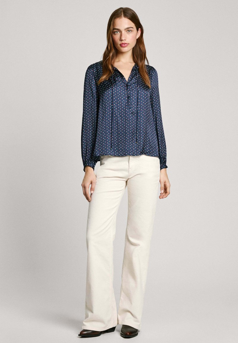 Blusa navy de tejido ligero y brillante, con un patrón de puntos, botones en la parte delantera y mangas largas; combinada con pantalones cremosos de campana.