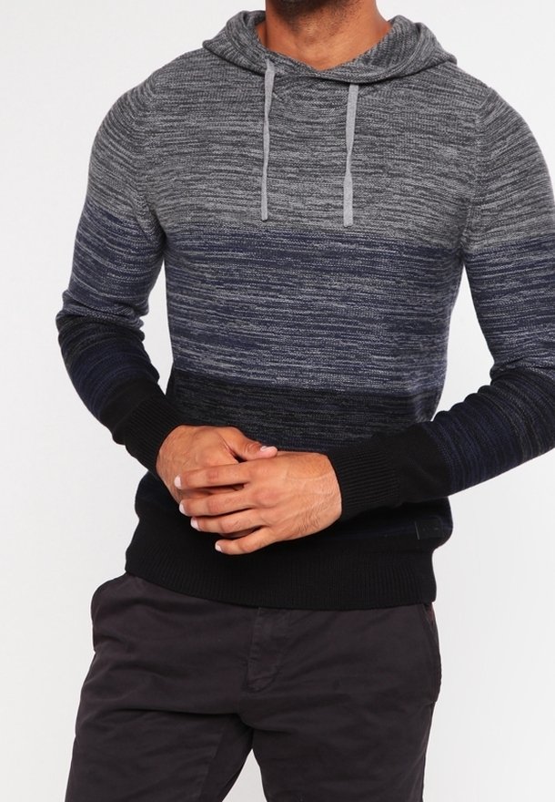 Man in een gradient gebreide hoodie in grijstinten, marineblauw en zwart, met koordjes, en donkere broek, met handen op de taille geklemd.