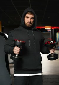 Felpa nera con cappuccio e cordino, che tiene due manubri neri con accenti rossi. Lo sfondo presenta attrezzature da palestra e un'illuminazione soffusa.