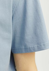 Camisa de algodón de manga corta azul claro con una textura suave, con costuras sencillas a lo largo de los bordes y un ajuste holgado en la zona de los brazos.