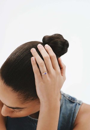 Anillo de oro con una piedra púrpura, usado en una mano que toca el cabello. El acabado suave, el diseño minimalista y la forma redonda enfatizan la gema.