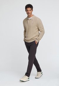 Beżowy sweter z grubym splotem i kołnierzem, zestawiony z ciemnymi spodniami i kremowymi trampkami z zielonymi akcentami. Gładka dzianina, casualowy design.