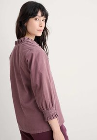 Ilgų rankovių bluzonas iš švelnaus melsvo audinio, su raukšlėtu apykaklė ir surinktais rankogaliais, suteikiančiais papildomos tekstūros.