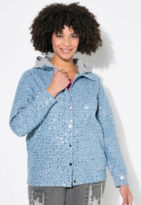 Veste en sequins bleu clair avec une capuche grise, dotée de boutons-pression argentés, d'une surface texturée et d'une coupe décontractée. Portée avec un pantalon gris.