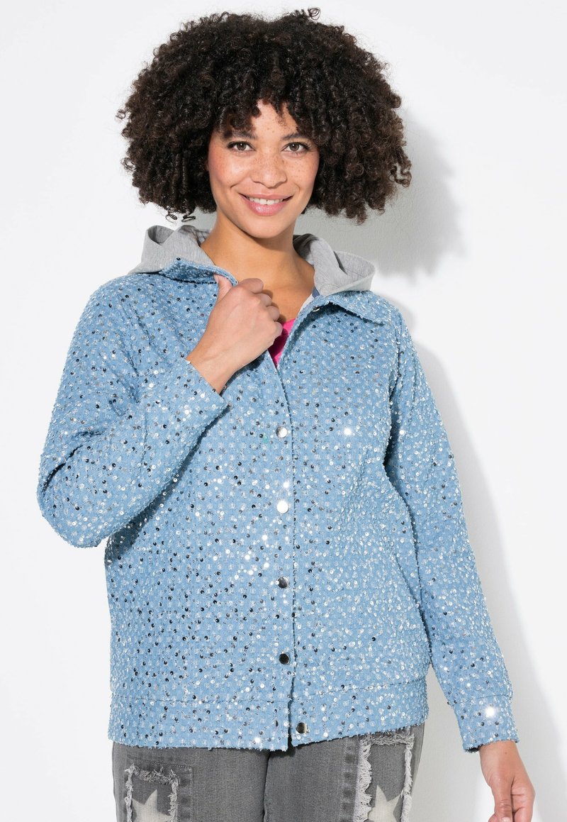 Veste en sequins bleu clair avec une capuche grise, dotée de boutons-pression argentés, d'une surface texturée et d'une coupe décontractée. Portée avec un pantalon gris.
