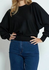 Musta ylipitkä neule, jossa on ribbireunukset ja hihansuut, yhdistettyinä tiukkoihin tummansinisiin farkkuihin. Materiaali näyttää pehmeältä ja malliltaan rento.