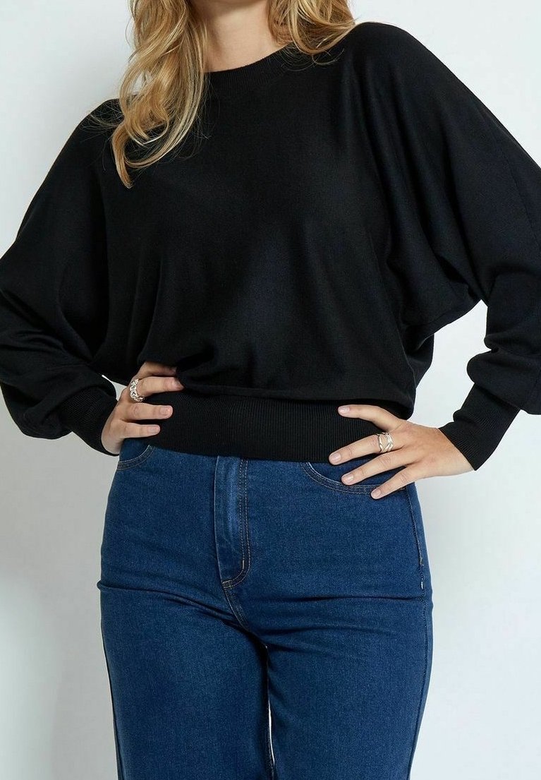 Musta ylipitkä neule, jossa on ribbireunukset ja hihansuut, yhdistettyinä tiukkoihin tummansinisiin farkkuihin. Materiaali näyttää pehmeältä ja malliltaan rento.