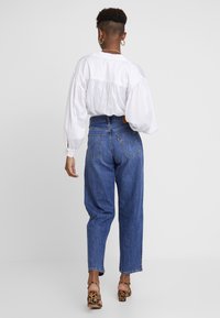 Valkoinen pallohiha-pusero, jossa kerätty selkäosa; siniset korkeavyötäröiset denimfarkut; eläinkuosiset korkokengät; yksinkertaiset kultaiset rinkkorenkaat.