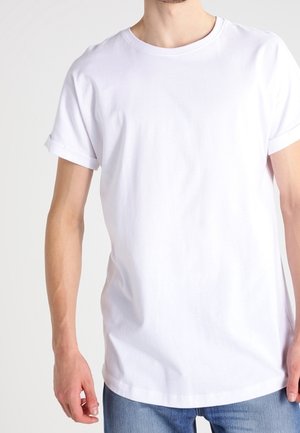 Mężczyzna ubrany w prosty biały t-shirt z krótkim rękawem i niebieskie dżinsy z podwiniętymi nogawkami, stojący na białym tle.