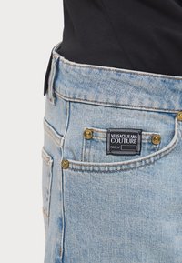 Lyseblå denimjeans med en gullknapp og sømdetaljer. Versace Jeans Couture-merket synlig på baklommen. Klassisk design.