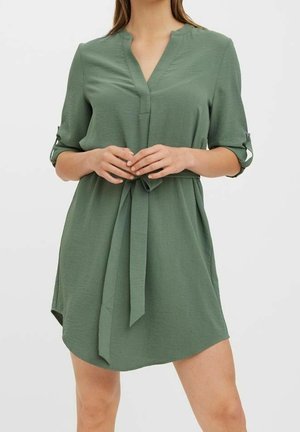 Frau trägt ein olivgrünes Kurzarmkleid mit V-Ausschnitt und Stoffgürtel, der in der Taille gebunden ist, und steht vor einem einfarbigen weißen Hintergrund.