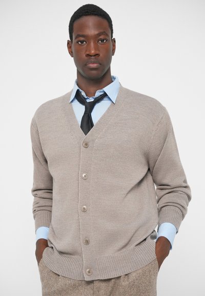 Νεαρός άνδρας που φοράει μπεζ πλεκτό καμπάνα πάνω από ανοιχτό μπλε πουκάμισο με μαύρη γραβάτα, με τα χέρια στις τσέπες, ουδέτερο φόντο.