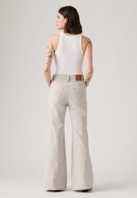 Jeans bege em estilo flare com cintura alta, apresentando bolsos traseiros e uma etiqueta de couro castanha na cintura, combinados com um top justo branco.