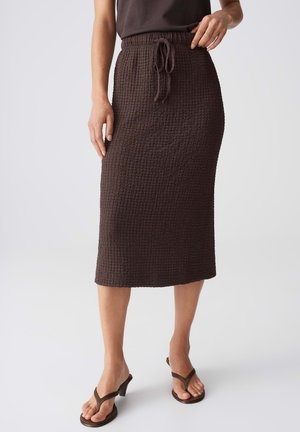 Donna che indossa una gonna midi marrone scuro strutturata con coulisse in vita, abbinata a sandali con tacco marrone scuro su uno sfondo semplice.
