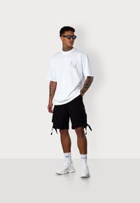 White cotton t-shirt with multicolored logo on the chest, paired with black cargo shorts featuring pockets and ties, and white sneakers.