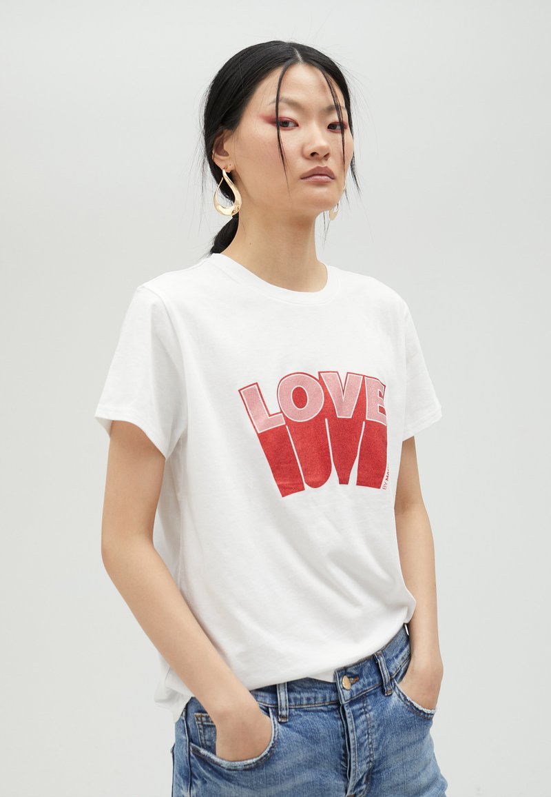 Camiseta de algodón blanca con un atrevido estampado gráfico rojo de "LOVE", de mangas cortas, con un corte relajado, combinada con pantalones cortos de mezclilla azul.