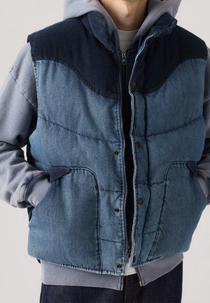 Personne portant une veste matelassée bleue avec un empiècement en velours côtelé sur un sweat à capuche gris, mains dans les poches de la veste, sur un fond uni.
