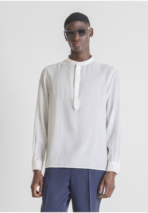 Homme portant des lunettes de soleil sombres, une chemise blanche à manches longues avec col mandarin et un pantalon bleu marine sur un fond clair uni.