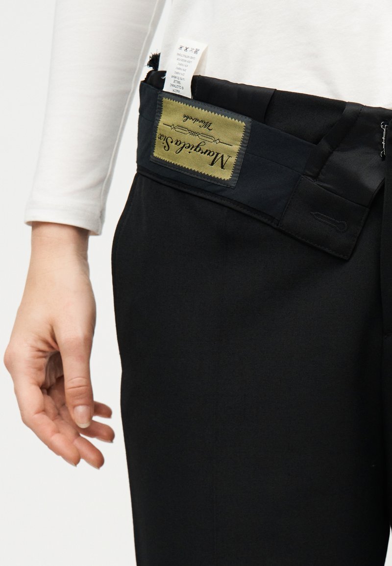 Gros plan sur une personne portant un pantalon noir avec la ceinture retournée, montrant une étiquette en tissu verte et noire ainsi qu'une étiquette d'entretien.