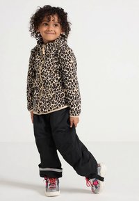 Veste polaire à imprimé léopard beige avec fermeture éclair à l'avant, associée à un pantalon ample noir. Les baskets présentent des accents métalliques et des lacets colorés.