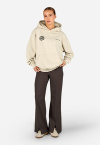 béžová mikina s kapucňou a predným vreckom s logom; skombinovaná s tmavohnedými nohavicami s rozparkom na leme a svetlými topánkami.