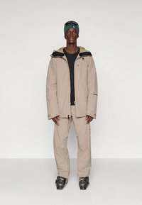 Beige waterdichte jas met een zwarte voering, gecombineerd met bijpassende beige broek en zwarte skibotten. Het model draagt ook een zwart shirt en een groene muts.