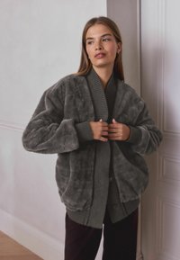 Šedá umělá kožená bunda s texturovaným povrchem, žebrovaným límcem a rukávy. Volný střih s bočními kapsami. Nositá přes tmavý top, na pozadí světlé zdi.