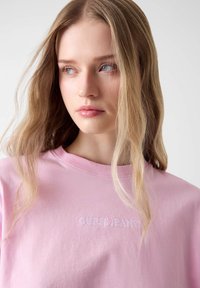 Różowy bawełniany t-shirt z okrągłym dekoltem i haftowanym logo "GUESS JEANS" na piersi, o miękkiej teksturze i luźnym kroju.