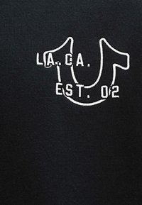 T-shirt en coton noir avec un graphique brodé blanc portant "LA. CA." et "EST. 02" ; le design comprend une grande forme en "U" avec des accents circulaires.