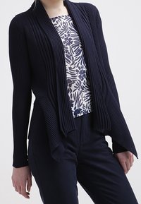 Mujer vistiendo un cárdigan oscuro con canalé y abertura frontal sobre una blusa blanca con estampado floral en azul marino y pantalones oscuros ajustados.