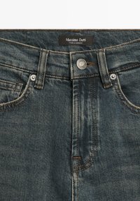 Gros plan sur la ceinture d'un jean en denim bleu avec bouton, passants de ceinture, poches avant et étiquette « Massimo Dutti Fabriqué au Maroc ».