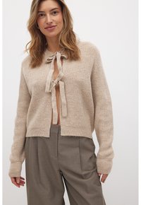 Cardigan beige in maglia con nodo frontale, texture a coste e maniche lunghe. Abbinato a pantaloni grigi a vita alta.