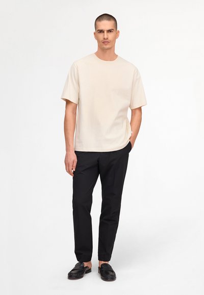 Uomo in piedi che indossa una T-shirt beige a maniche corte, pantaloni neri slim fit e mocassini neri slip-on su uno sfondo bianco.
