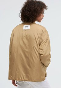 Beige Oversized-Jacke mit glatter, glänzender Textur, die ein Etikett auf der Rückseite hat. Sie hat einen abgerundeten Saum und eine lockere Passform.