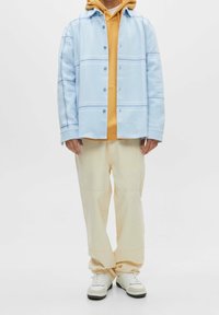 Camicia azzurro chiaro con righe orizzontali, sovrapposta a una felpa gialla, abbinata a pantaloni crema e sneakers bianche.