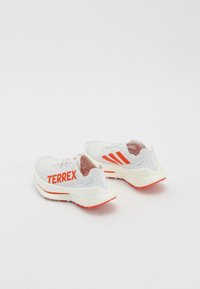 White athletic shoes with orange accents and "TERREX" printed on the side. Features a padded tongue, textured upper, and rubber sole.