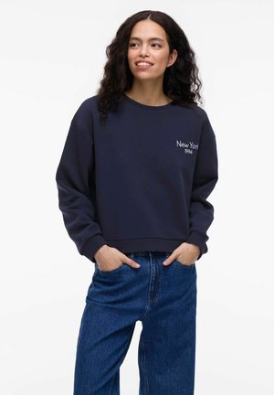 Donkerblauwe sweatshirt met ribbelboord aan de nek en mouwen, voorzien van witte borduring "New York 1994" op de voorkant. In combinatie met losse blauwe jeans.