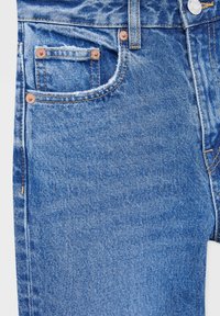 Gros plan sur un jean en denim bleu clair montrant la poche avant, la petite poche avec rivets métalliques, une passoire pour ceinture, un bouton et des coutures visibles.