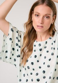 Luftige, leichte Bluse in Creme mit einem schwarzen und tealfarbenen Punktmuster, ausgestattet mit lockeren Ärmeln und einem runden Ausschnitt. Sanfte Stofftextur.