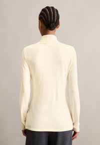 Long-sleeve, cream turtleneck shirt made of soft fabric. Features a fitted design with a high neckline and textured detailing on the back.