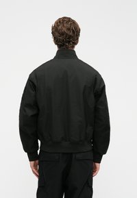 Jachetă neagră tip bomber, cu guler înalt, manșete și tiv ribbed, fabricată dintr-un material neted și ușor. Prezintă o croială lejeră și un design minimalist.