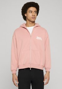 Lys rosa hettegenser med glidelås, laget av mykt stoff, med frontlommer og hvit trykt tekst på venstre brystområde. Lange ermer med ribbestrikkede mansjetter.