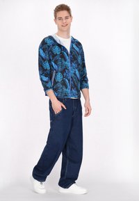 Blauwe hoodie met rits en tropisch loofpatroon, witte binnenvoering, gecombineerd met losse donkere spijkerbroek en witte sneakers.