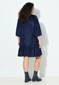 Nainen, jolla on kihara hius, pukeutuneena tummansiniseen polvipituiseen mekkoon, jossa on 3/4-hihat ja tummansiniset nilkkurit, seisoo valkoista taustaa vasten.