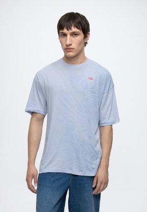 Junger Mann mit kurzen dunklen Haaren, der ein hellblaues Diesel-T-Shirt und blaue Jeans trägt, vor einem schlichten weißen Hintergrund.