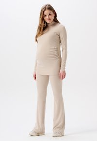 Umstandsoutfit aus leicht beigem, geripptem Stoff, bestehend aus einem Langarmoberteil mit hohem Kragen und einer engen Hose, die für Komfort und Dehnbarkeit konzipiert ist.