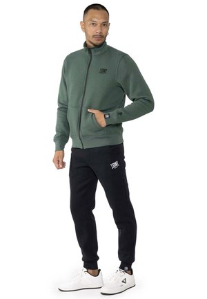 Un homme porte une veste de survêtement verte zippée, un pantalon de jogging noir et des baskets blanches sur un fond blanc uni.