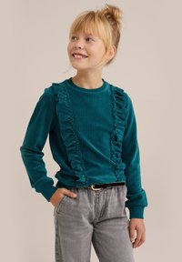 Tirkizna kordurojeva srajca z dolgimi rokavi in volančki ob sprednjih straneh, rebrasta tekstura in okrogla ovratnika, v kombinaciji s sivimi jeansami.